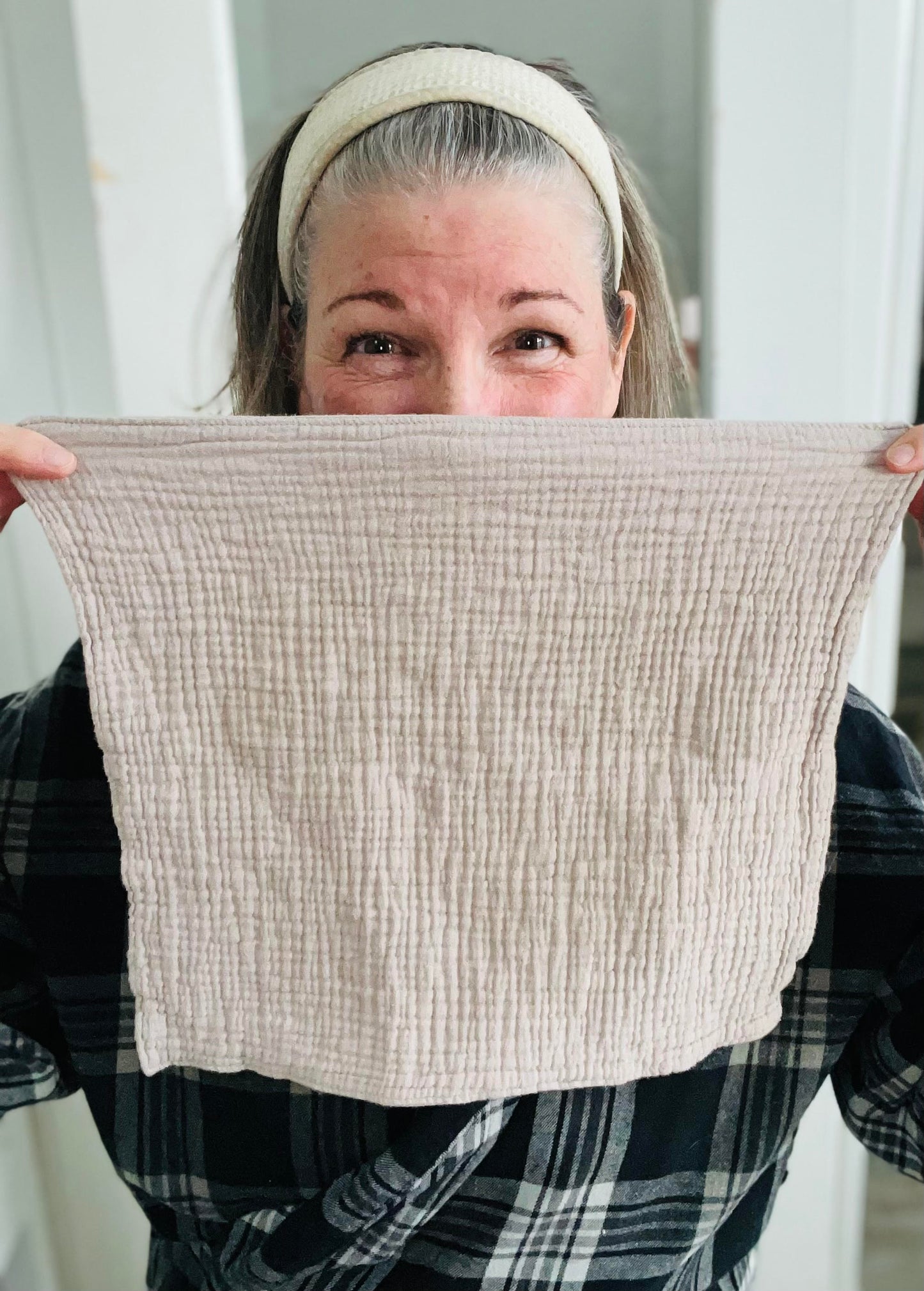 A picture of a woman (me!) with her hair pulled back with a hairband. She smiles at the camera and holds a fawn (light taupe) coloured face cloth in front of her face, covering her nose to her mid-chest. The wavy and pillowy texture cleansing cloth is on display.