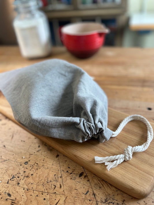 A hand-sewn linen bread bag holds a loaf. The recycled cotton cord is drawn tight to protect the bread. It lays on a wooden board with a red bowl in the background.
