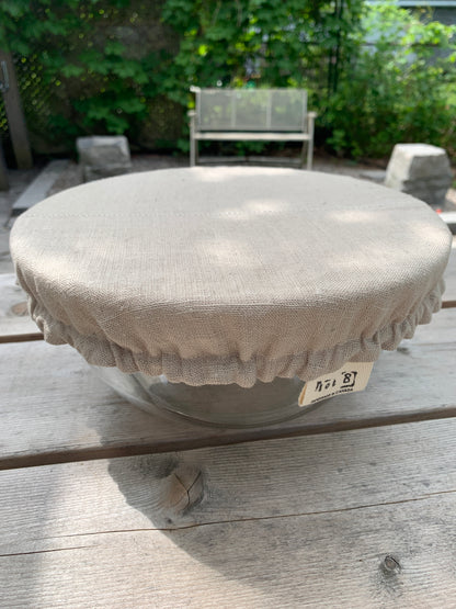 A bowl covered with a linen bowl cover sits on a picnic table. A bench in front of a wall of ivy is blurred in the background