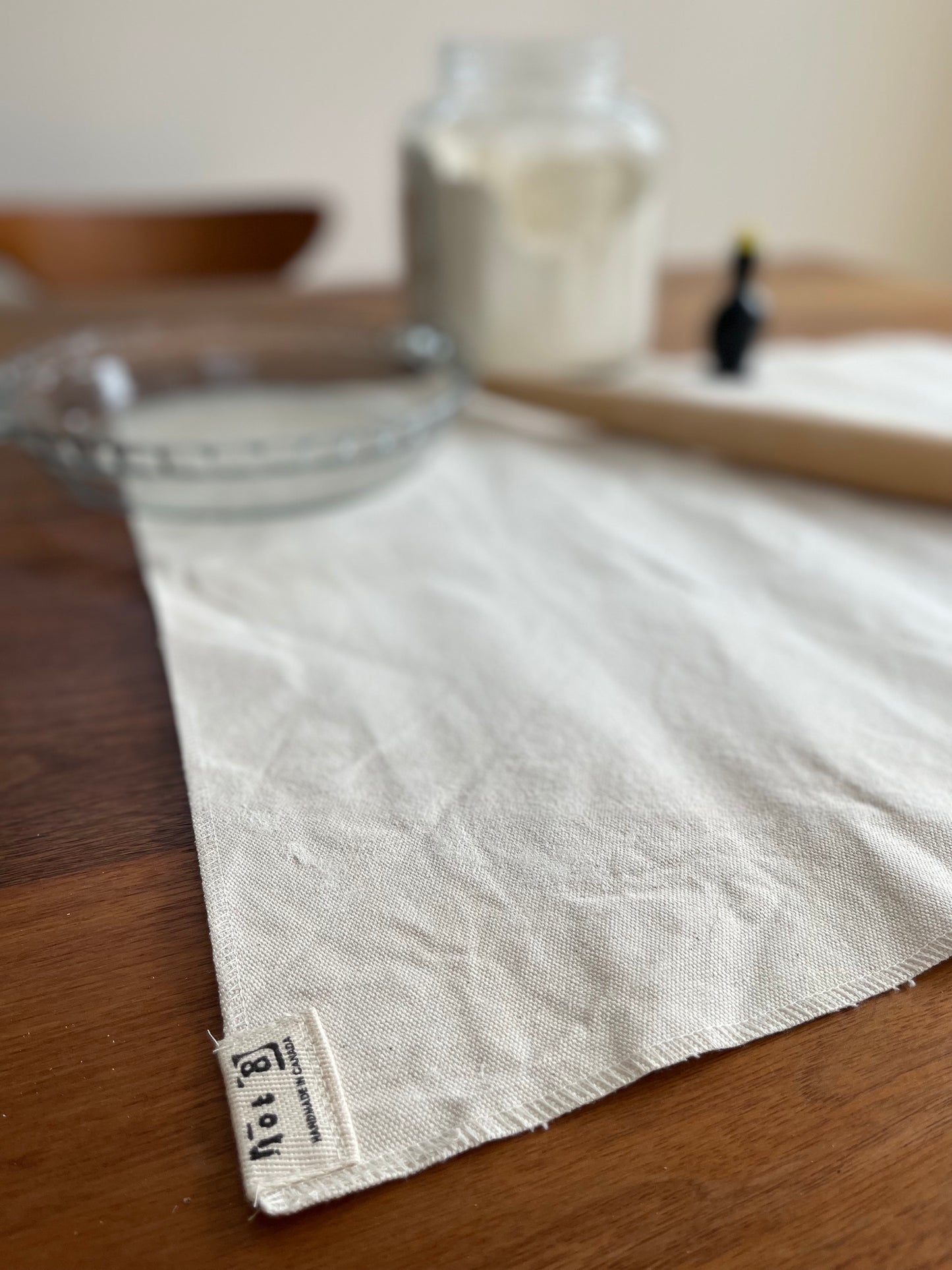 A pastry cloth showing a lot8 tag in the foreground shows an empty pie plate, rolling pin, blackbird pie vent and jar of flour in the background.