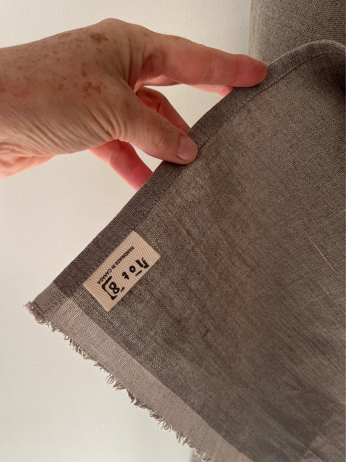 A hand hold up the corner of the natural linen tea towel revealing the cotton twill lot8 tag sewn into the bottom corner
