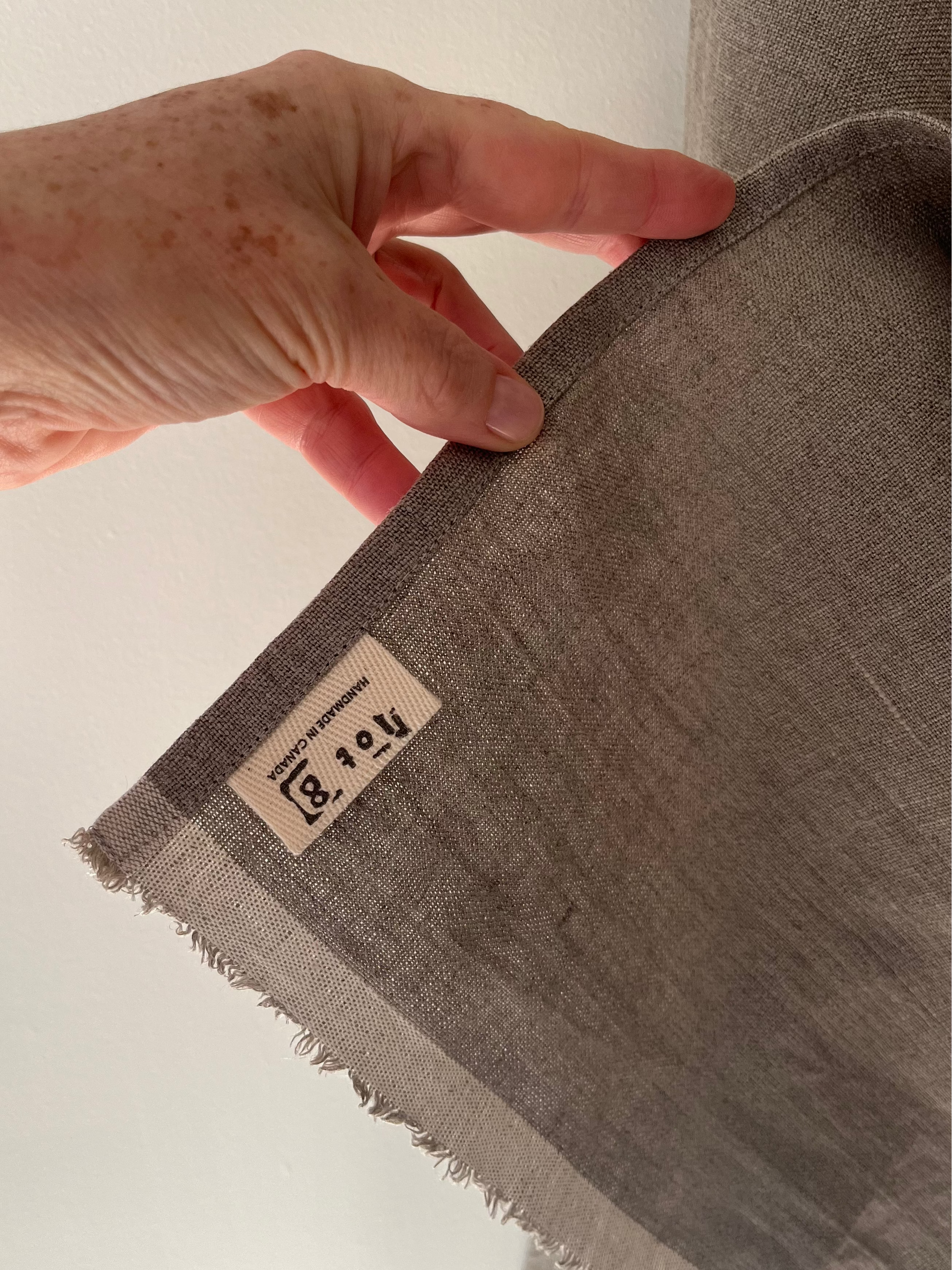 A hand hold up the corner of the natural linen tea towel revealing the cotton twill lot8 tag sewn into the bottom corner