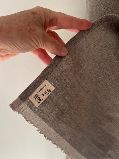 A hand hold up the corner of the natural linen tea towel revealing the cotton twill lot8 tag sewn into the bottom corner