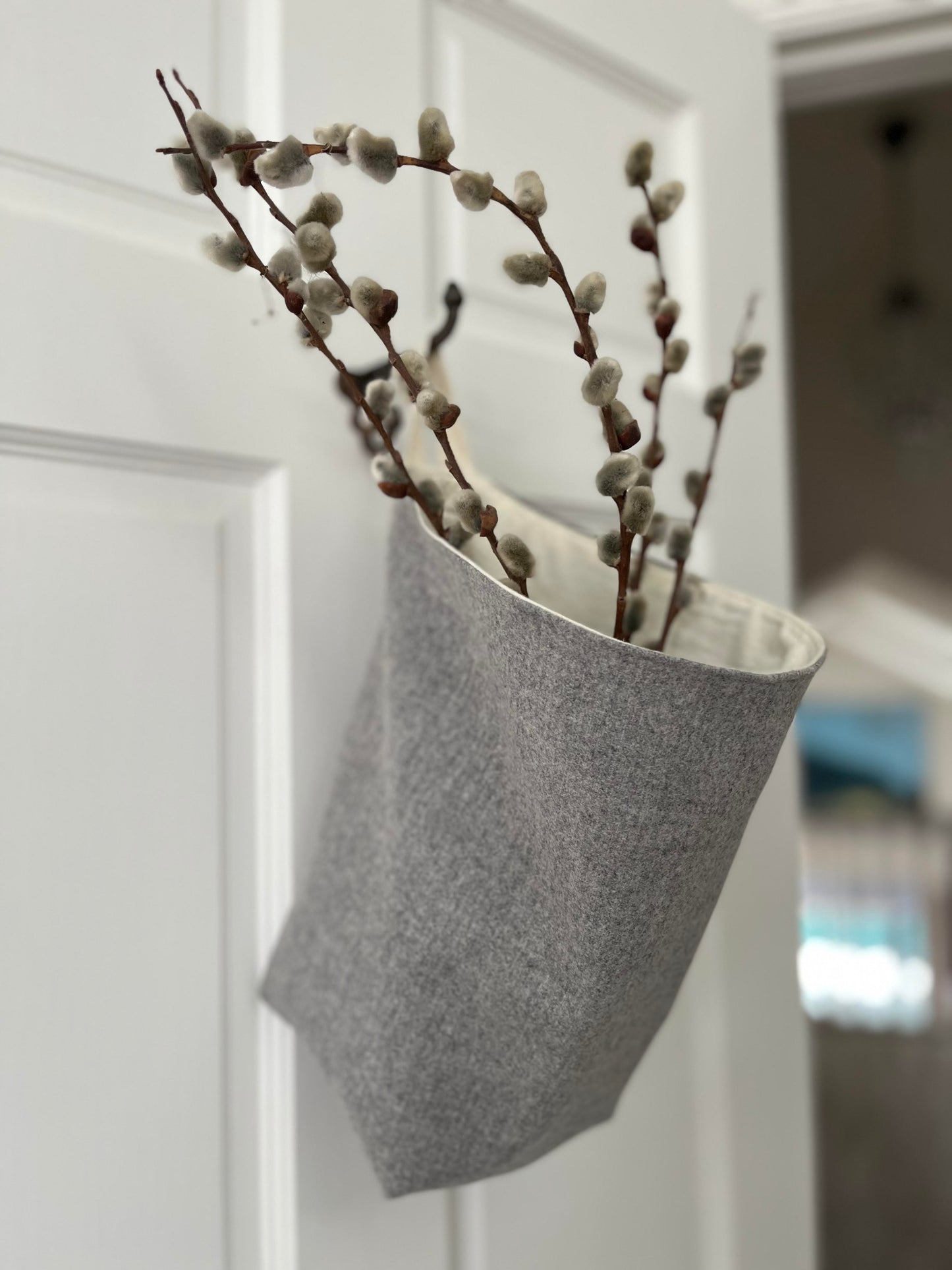 A wool sack lined with ivory flannel hangs from a hook on a white bedroom door. It is filled with soft grey pussy willows.