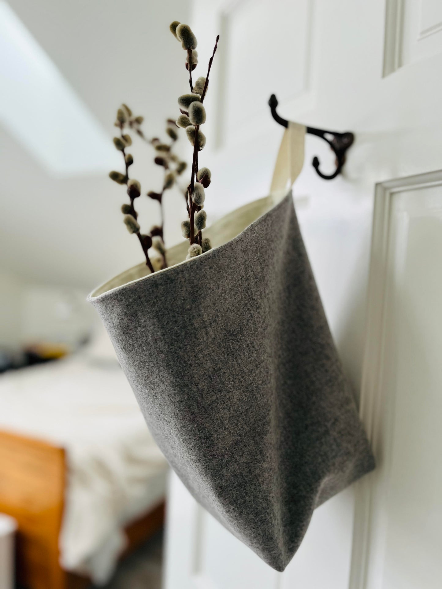The side view of the all purpose sack filled with pussy willows hanging from a hook on  the bedroom door. The bed with a white duvet is blurred in the backgrounc.